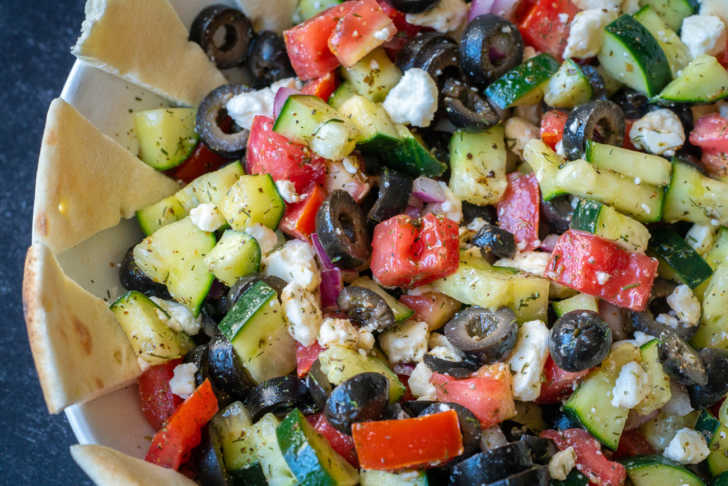 A bowl of vegetables including tomatoes, cucumbers, olives, and black olives.
